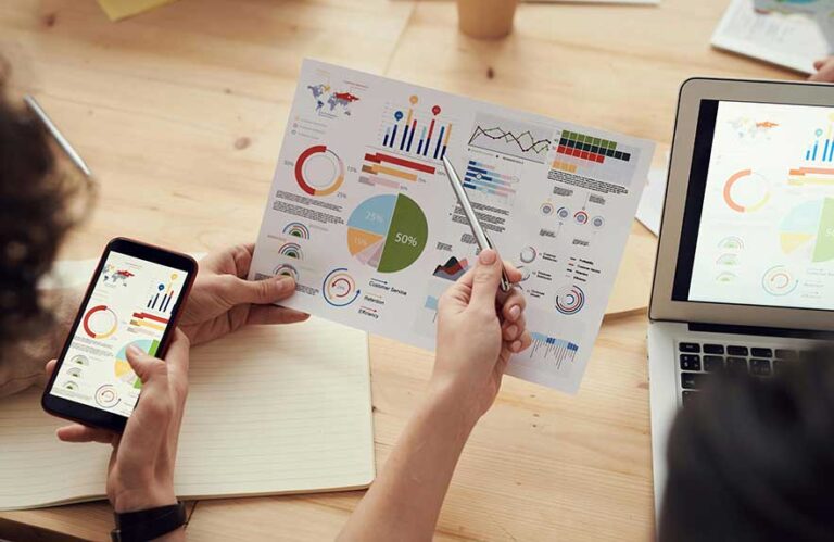 Hands comparing charts on phone, paper, and laptop, illustrating free vs paid productivity tools in a modern office setup